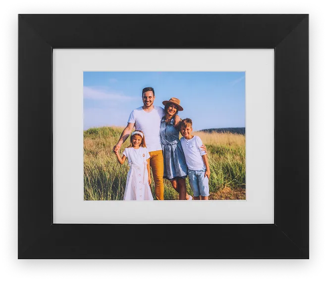 Framed family photo of two parents and two children standing in a grassy field at golden hour, in a black wood frame.