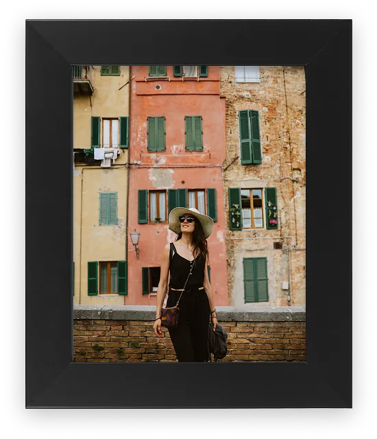 Framed portrait of a woman in a sun hat and black outfit standing in front of colorful Italian buildings, in a black wood frame.